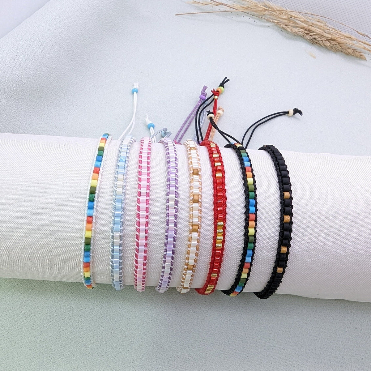Eight thin beaded macramé bracelets in rainbow, blue, pink, purple, cream, red and black, stacked on a white roll