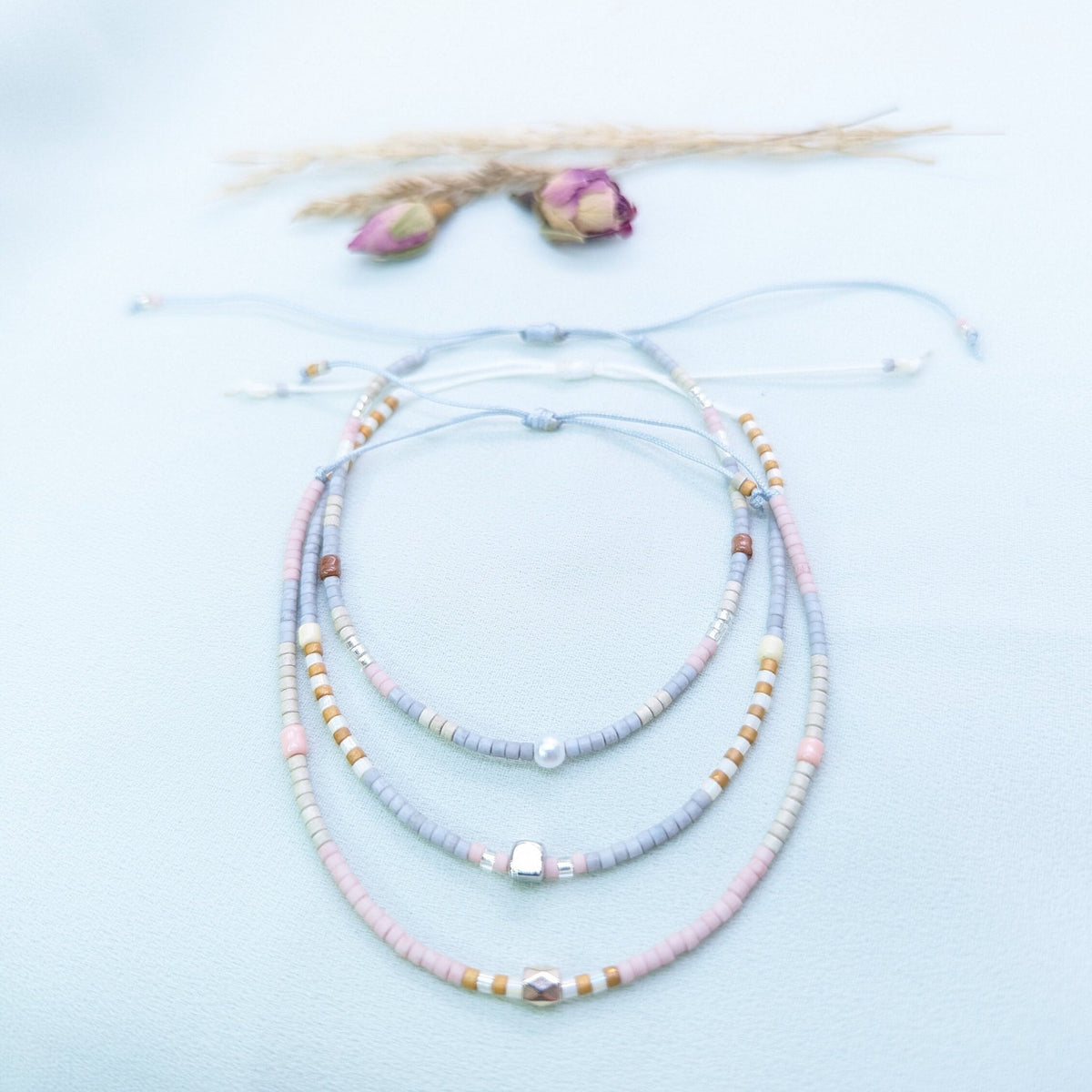 Set of three grey, white and pastel seed bead bracelets with adjustable cords, beside dried rosebuds