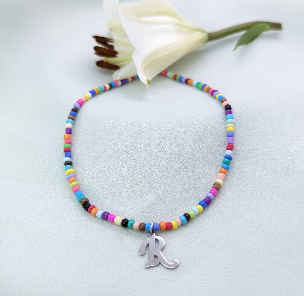 Multicoloured seed bead necklace with a silver script initial R pendant, laid on white with a lily