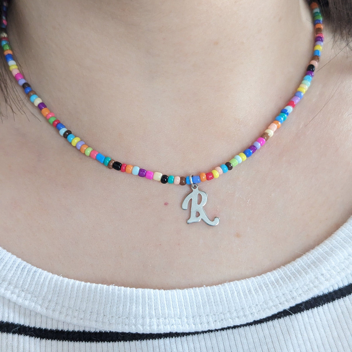 Rainbow seed bead necklace with a silver script letter R charm worn on the neck with a striped top