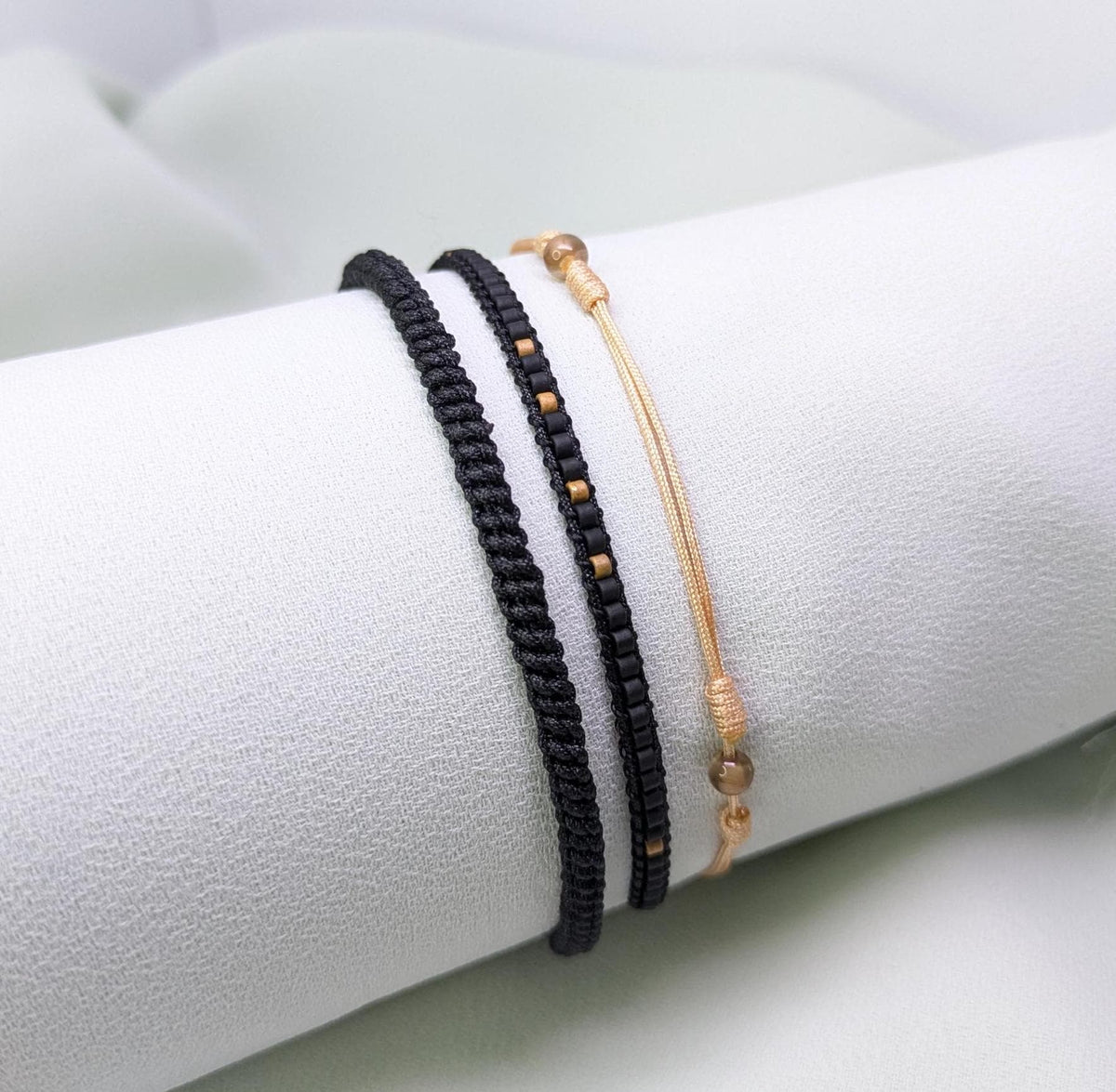 Three black and caramel woven and string bracelets with gold bead accents, on a white roll