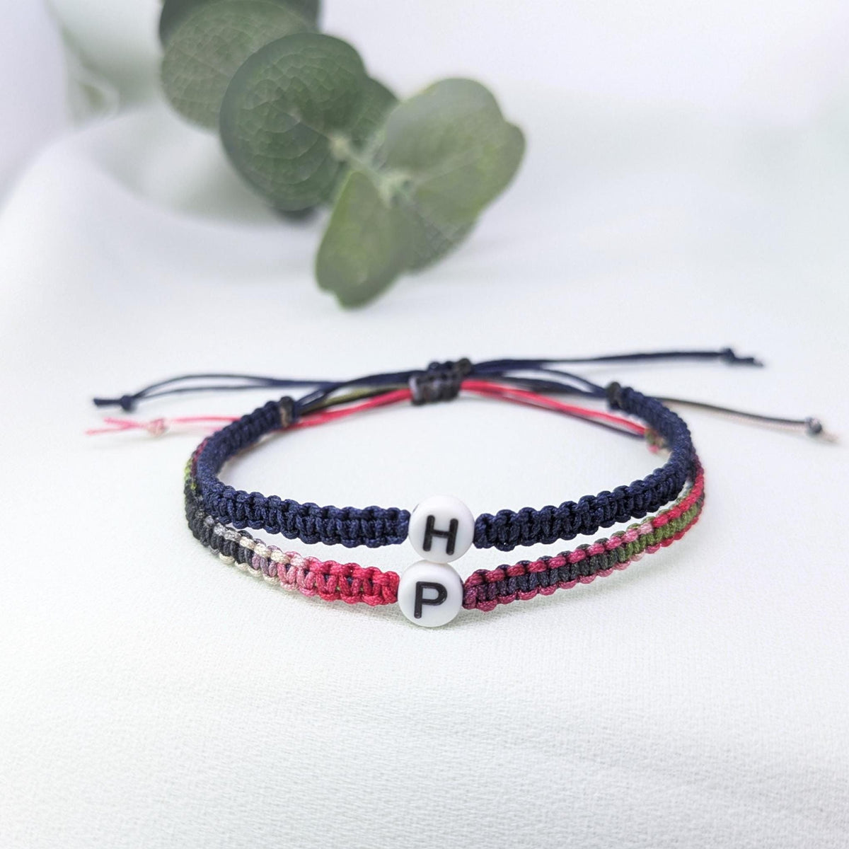 Navy and pink woven couple bracelets with white initial beads H and P, laid together on white